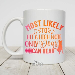 May include: White ceramic mug with a handle. The mug features the text "MOST LIKELY TO HIT A HIGH NOTE ONLY DOGS CAN HEAR" in pink and orange. A silhouette of a dog is also on the mug.