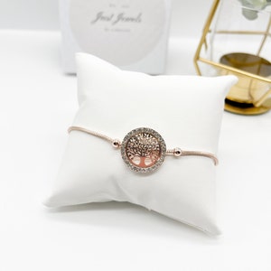 May include: A rose gold bracelet with a tree of life charm. The charm is encrusted with clear rhinestones. The bracelet is on a white pillow.