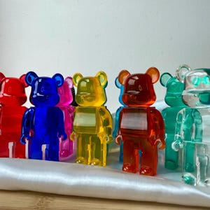 May include: Seven colorful, transparent, bear-shaped figurines. The bears are made of a clear plastic material and are in various colors, including red, blue, yellow, green, and clear. The bears are all standing on a white surface.