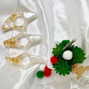 May include: Three white bookmark holders with gold flecks. The holders are shaped like a curved wing. The holders are on a white surface.