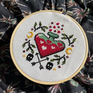 May include: A cross stitch hoop with a heart-shaped potion bottle with a green top and red liquid. The bottle is surrounded by a wreath of leaves and has a crescent moon on each side. There are two dice and two arrows crossing each other below the bottle.