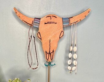 Può includere: Un porta gioielli in legno a forma di teschio di toro con una base marrone e un'asta in metallo dorato. Il supporto ha una fascia di motivo blu, bianco e marrone attorno al cranio. Ci sono diverse collane appese alle corna e al cranio.