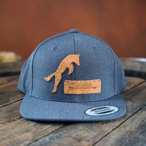 May include: A dark gray baseball cap with a brown leather patch featuring a jumping horse silhouette and the text "SPRITZSTOCK".