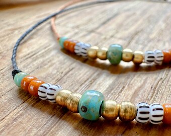 Può includere: Primo piano di una collana di perline con perline arancioni, bianche e dorate. La collana ha una perla turchese al centro.