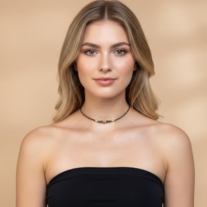 Pu&ograve; includere: Una collana girocollo marrone con un elemento decorativo centrale con perline rosse, verdi e dorate. La collana &egrave; indossata intorno al collo. Lo sfondo &egrave; di colore beige.