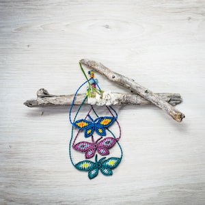 May include: Three colorful butterfly-shaped friendship bracelets made with knotted string, hanging from a piece of driftwood.