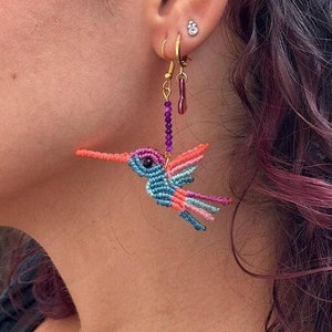 May include: A pair of dangle earrings featuring a colorful hummingbird made of thread. The hummingbird is blue, pink, orange, and purple. The earrings are hanging from gold hooks.
