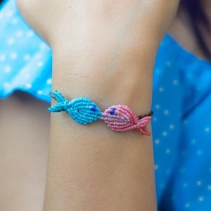 Op de afbeelding: Een vriendschapsarmband met twee visbedels, een blauwe en een roze. De armband is gemaakt met een knooptechniek en wordt om de pols gedragen.