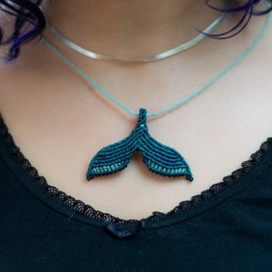 May include: A teal blue whale tail pendant necklace on a silver chain. The pendant is made of macrame and has a light blue and dark blue color gradient.