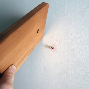 May include: A close-up of a wooden board being held up to a white wall with a screw already in place.