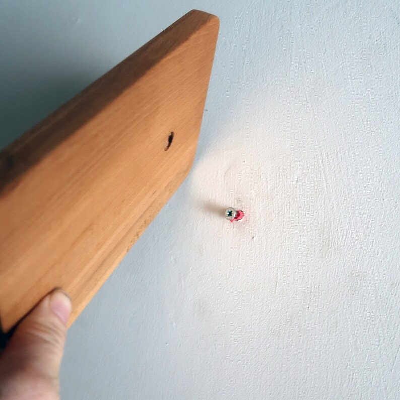 May include: A close-up of a wooden board being held up to a white wall with a screw already in place.