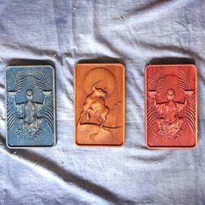 May include: Three wooden plaques with carved designs. The left plaque is blue and features a person holding scales. The center plaque is brown and features a bull. The right plaque is red and features a person holding scales.