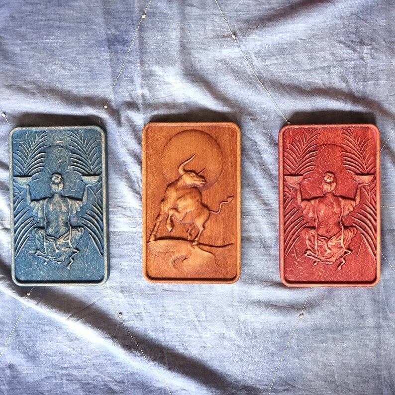 May include: Three wooden plaques with carved designs. The left plaque is blue and features a person holding scales. The center plaque is brown and features a bull. The right plaque is red and features a person holding scales.