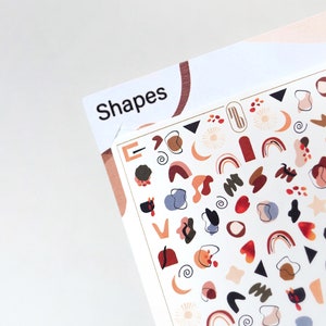May include: A sheet of nail art stickers with the word "Shapes" at the top. The stickers feature a variety of abstract shapes in shades of brown, orange, red, blue, and green.