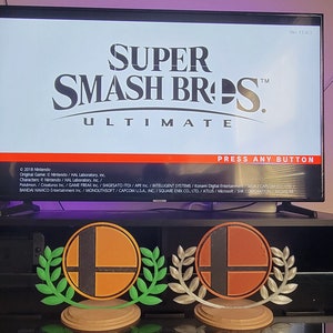 Puede incluir: Dos trofeos de Super Smash Bros. Ultimate, uno con una base verde y otro con una base plateada. Los trofeos presentan el logotipo de Super Smash Bros. con una corona de laurel alrededor. Los trofeos están colocados en un estante negro frente a una pantalla de televisión que muestra la pantalla de título de Super Smash Bros. Ultimate.