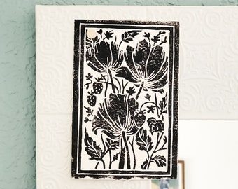 Pode incluir: Obra de arte com estampa floral em preto e branco com flores e morangos estilizados. A obra de arte é montada em um fundo branco com um padrão decorativo em relevo. Um pequeno fecho redondo branco é visível.