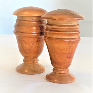 May include: Two wooden salt and pepper shakers with a turned wood design. The shakers are a light brown color with a reddish hue.