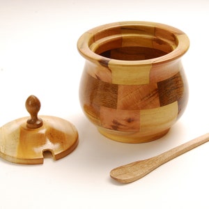 May include: A wooden sugar bowl with a lid and a small wooden spoon. The bowl is made of different types of wood, creating a patchwork pattern.