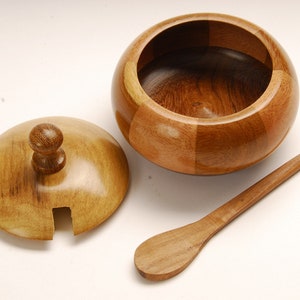 May include: A wooden sugar bowl with a lid and a small wooden spoon. The bowl is round with a lid that has a small knob on top. The spoon is long and thin. The bowl and spoon are made of light and dark brown wood.