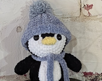 May include: A handmade crochet penguin with a light blue hat and scarf. The penguin has a white belly, black wings, and a yellow beak. The eyes are black. The penguin is a soft, plush toy.