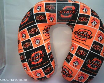 May include: Orange and black travel neck pillow featuring a repeating pattern of the Oklahoma State University logo, the letters "OSU", and a cartoon cowboy. The pillow is designed for comfort and support during travel.