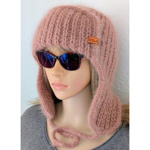 Könnte beinhalten: Eine staubrosa gestrickte Trappermütze mit Ohrenklappen und einem Kordelverschluss. Die Mütze hat ein kleines rechteckiges Etikett mit Text. Das Model trägt eine schwarze Sonnenbrille. Die Mütze ist aus weichem, flauschigem Material.