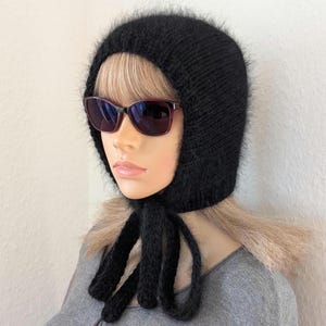 May include: A black knitted hood with long ties, worn with dark sunglasses. The hood is made of a fuzzy, textured yarn. The ties hang down the front. The hood covers the head and neck, leaving the face exposed.