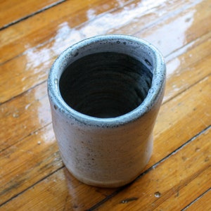 Könnte beinhalten: Eine weiße Keramikvase mit blauer Glasur und einer rauen, strukturierten Oberfläche. Die Vase ist leer und hat eine runde Öffnung oben.