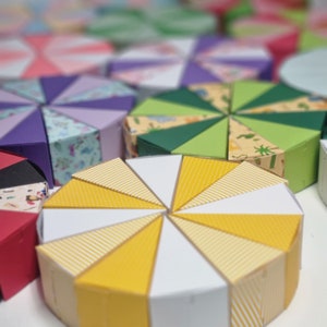 Pode incluir: Uma variedade de caixas de bolo de papel coloridas e estampadas, em diferentes formas e tamanhos. Algumas caixas estão abertas, revelando um bolo listrado amarelo e branco no interior.
