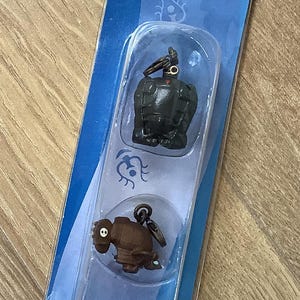 May include: A blue package containing three miniature keychains. The keychains feature characters from the movie "Castle in the Sky". The top keychain is a dark gray robot, the middle is a brown creature, and the bottom is a green gem.