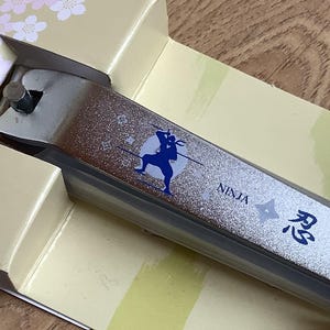 Könnte beinhalten: Silberne Nagelknipser mit einer blauen Ninja-Grafik und japanischem Text, verpackt in einer beige-blauen Schachtel. Die Knipser haben eine strukturierte Oberfläche und werden auf einer Holzoberfläche präsentiert.