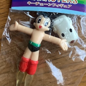 May include: A keychain featuring a Tezuka Osamu character figure. The figure is of a cartoon character with black shorts, red boots, and a green belt. The keychain also includes a white, ghost-like figure. The packaging has the text "TEZUKA OSAMU CHARACTERS".