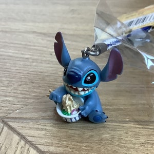 Peut inclure: Figurine bleue de Stitch avec de grandes oreilles et un large sourire, tenant une petite assiette de nourriture. La figurine est attachée à un porte-clés et est partiellement enfermée dans un emballage plastique transparent. Le thème général est Disney.