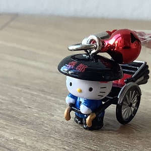 Peut inclure: Figurine Hello Kitty vêtue d'une tenue japonaise traditionnelle tirant un pousse-pousse miniature. La figurine porte un chapeau noir avec du texte japonais et une tenue bleue et blanche. Un ornement rouge est suspendu au-dessus du pousse-pousse.