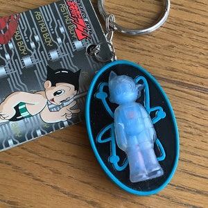May include: Astro Boy keychain featuring a translucent blue figure within a black oval with a blue border. Includes a metal keyring and a card with Astro Boy graphics and text.