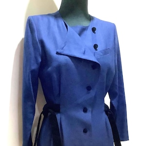 May include: A long-sleeved, knee-length blue dress with a black velvet belt. The dress features a button-down front, a unique collar design, and a small pocket. The dress is displayed on a mannequin.