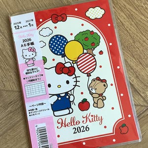 Puede incluir: Agenda Hello Kitty 2026 A6 con borde rojo. La portada presenta a Hello Kitty sosteniendo globos, un oso de peluche y un manzano. La agenda incluye texto japonés y un código de barras.