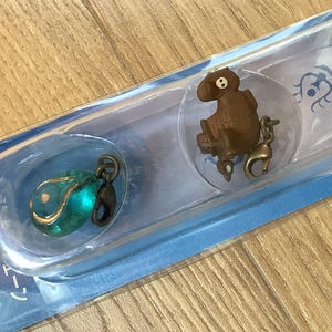 May include: A package containing three small charms. One is a teal orb, another is a brown figure, and the last is a dark green object. Each charm has a metal clasp for attachment. The packaging has Japanese text.