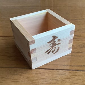 May include: A small, square wooden box with dovetail joints and a Japanese character on one side. The box is made of light-colored wood and sits on a wooden surface. The character is in a dark brown color.