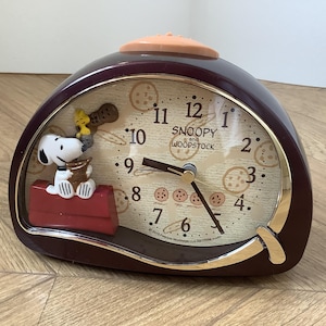 Peut inclure: Un réveil analogique semi-circulaire marron foncé représentant Snoopy et Woodstock sur une niche rouge. Le cadran affiche le texte Snoopy and Woodstock, des chiffres noirs et un motif de biscuits. Il possède des aiguilles dorées.