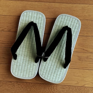 Könnte beinhalten: Ein Paar traditionelle japanische Geta-Sandalen. Die Sandalen haben ein hellgrünes Fußbett aus gewebtem Stroh mit weißem Rand und schwarze Stoffriemen. Die Geta liegen auf einem Holzboden.