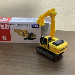 May include: A yellow and black Komatsu excavator toy with a movable arm, next to its box. The box features the text "KOMATSU EXCAVATOR PC200-10" and "SCALE 1/122". The toy is a miniature replica of a construction vehicle.
