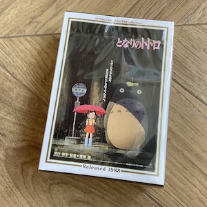 May include: A Studio Ghibli poster collection featuring the film "My Neighbor Totoro." The poster shows a young girl with a red umbrella at a bus stop, with Totoro standing nearby. The poster is in a white frame and is labeled "Released 1988."