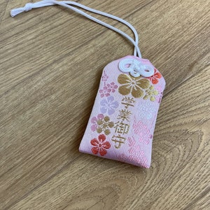 May include: A pink Japanese omamori charm with floral embroidery in gold, orange, and purple. The charm has Japanese text and a white cord. The charm is a traditional good luck item.