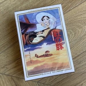 May include: A Studio Ghibli poster collection box featuring the movie "Porco Rosso." The artwork depicts a pig in a plane with a woman in a white hat. The poster has Japanese text and the release year 1992.