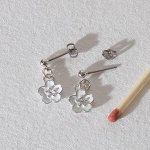 Op de afbeelding: Een paar zilverkleurige oorhangers met witte bloemvormige hangers. De oorbellen worden gepresenteerd met een lucifer op een wit oppervlak. De oorbellen hebben een stift en vlindersluiting.