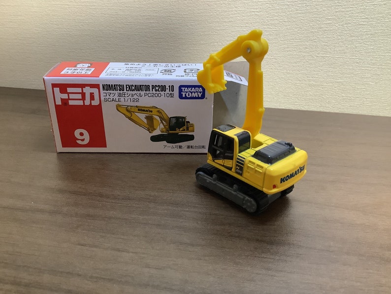 May include: A yellow and black Komatsu excavator toy with a movable arm, next to its box. The box features the text "KOMATSU EXCAVATOR PC200-10" and "SCALE 1/122". The toy is a miniature replica of a construction vehicle.