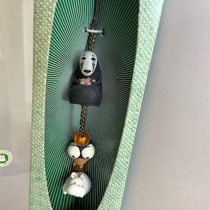 Pode incluir: Um ornamento decorativo suspenso com uma figura preta sem rosto, um cordão trançado castanho, uma conta âmbar translúcida, um par de olhos brancos arregalados e uma pequena figura branca semelhante a um rato, tudo dentro de uma caixa verde.