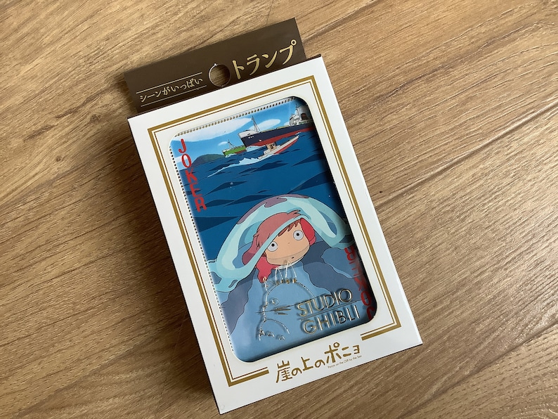 May include: A deck of playing cards in a white box with gold trim. The card face features a blue ocean scene with a ship and a character with pink hair under a blue blanket. The text includes JOKER, STUDIO GHIBLI, and Japanese characters.