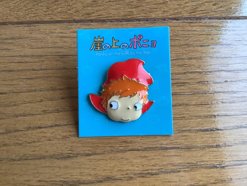 May include: A metal enamel pin featuring a cartoon character with orange hair and a red hood, attached to a bright blue backing card. The card displays the Japanese text 崖の上のポニョ and the English text Ponyo on the Cliff by the Sea.